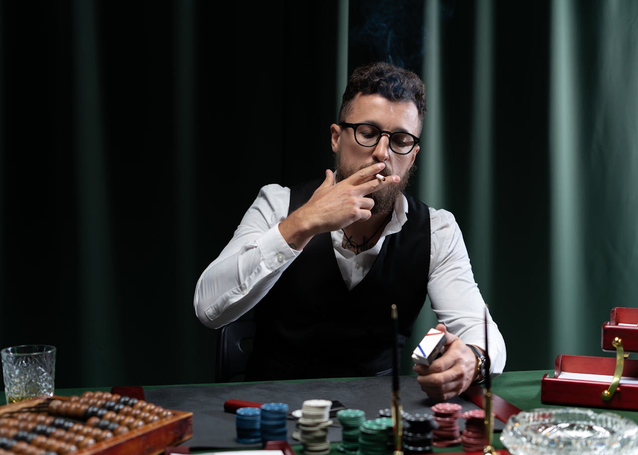 Man in glasses smoking at a poker table, surrounded by chips and cards, in a sophisticated indoor setting.